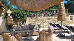 A sunlit outdoor seating area with wicker furniture, a stone wall, and a thatched umbrella. - La Bastide perched village in the Luberon (Ménerbes)