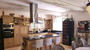 A modern kitchen with wooden cabinets, a central island with chairs, a built-in oven, and a stainless steel range hood. - La Bastide perched village in the Luberon (Ménerbes)