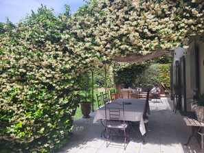 Een terras buiten met een tafel en stoelen, omringd door weelderig groen en bloeiende planten.