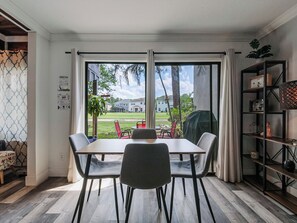 Un área de comedor con una mesa y sillas, ventanas amplias con cortinas, una estantería y vista a una casa y árboles.