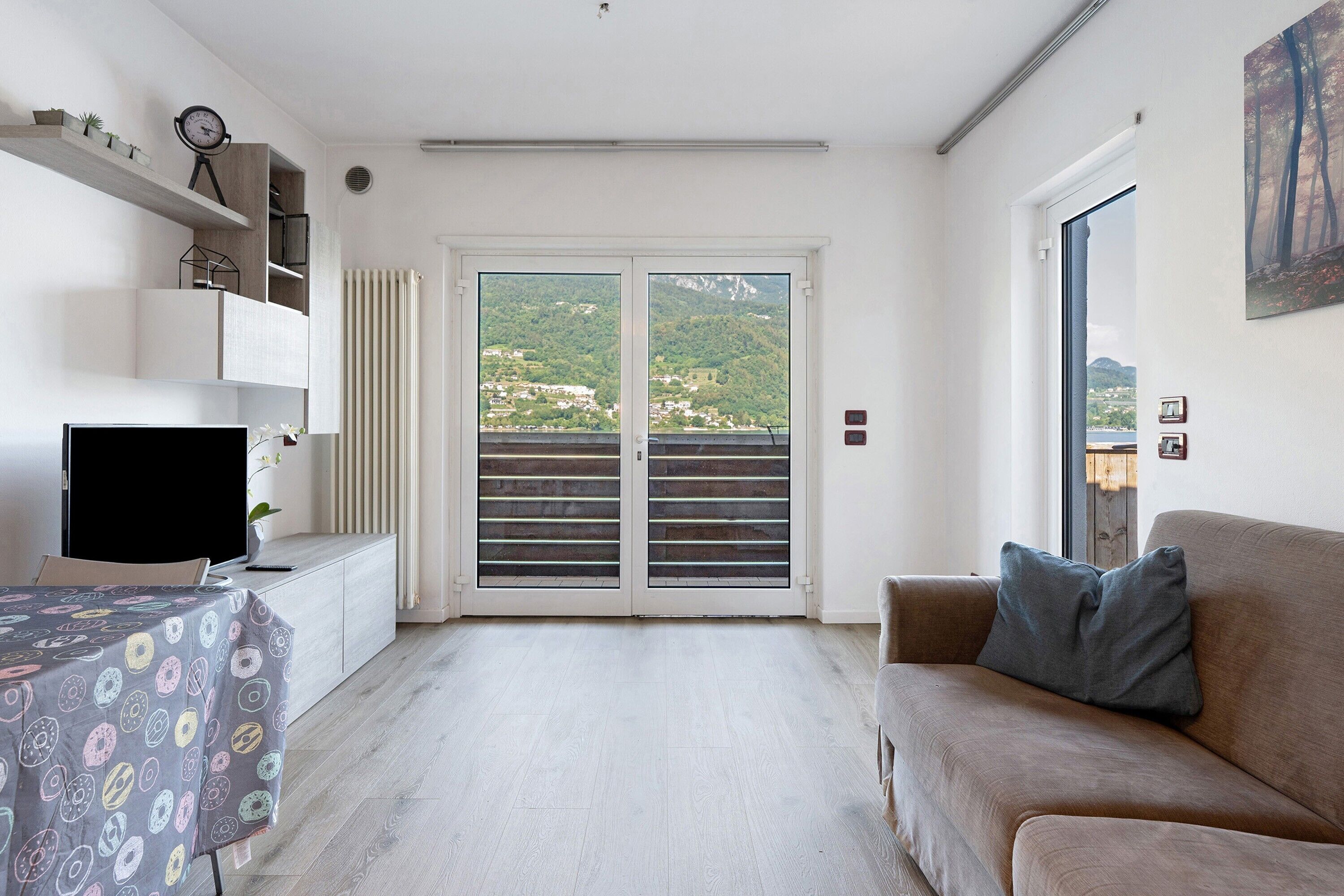 A modern living room with a sofa, a television, a table with a patterned tablecloth, a radiator, and a view of a green landscape through a sliding door.