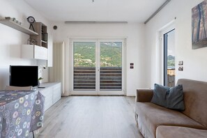 Ein modernes Wohnzimmer mit einer Couch, einem Fernseher, einem Tisch mit gemustertem Tischläufer, einer Heizung und Blick auf eine grüne Landschaft durch eine Schiebetür.