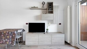 A modern living room with a flat-screen TV, a dining table with a donut-patterned tablecloth, and a potted orchid. - Apartment 'Valsugana D’incanto' with Lake View, Private Terrace and Shared Garden (Pergine Valsugana)