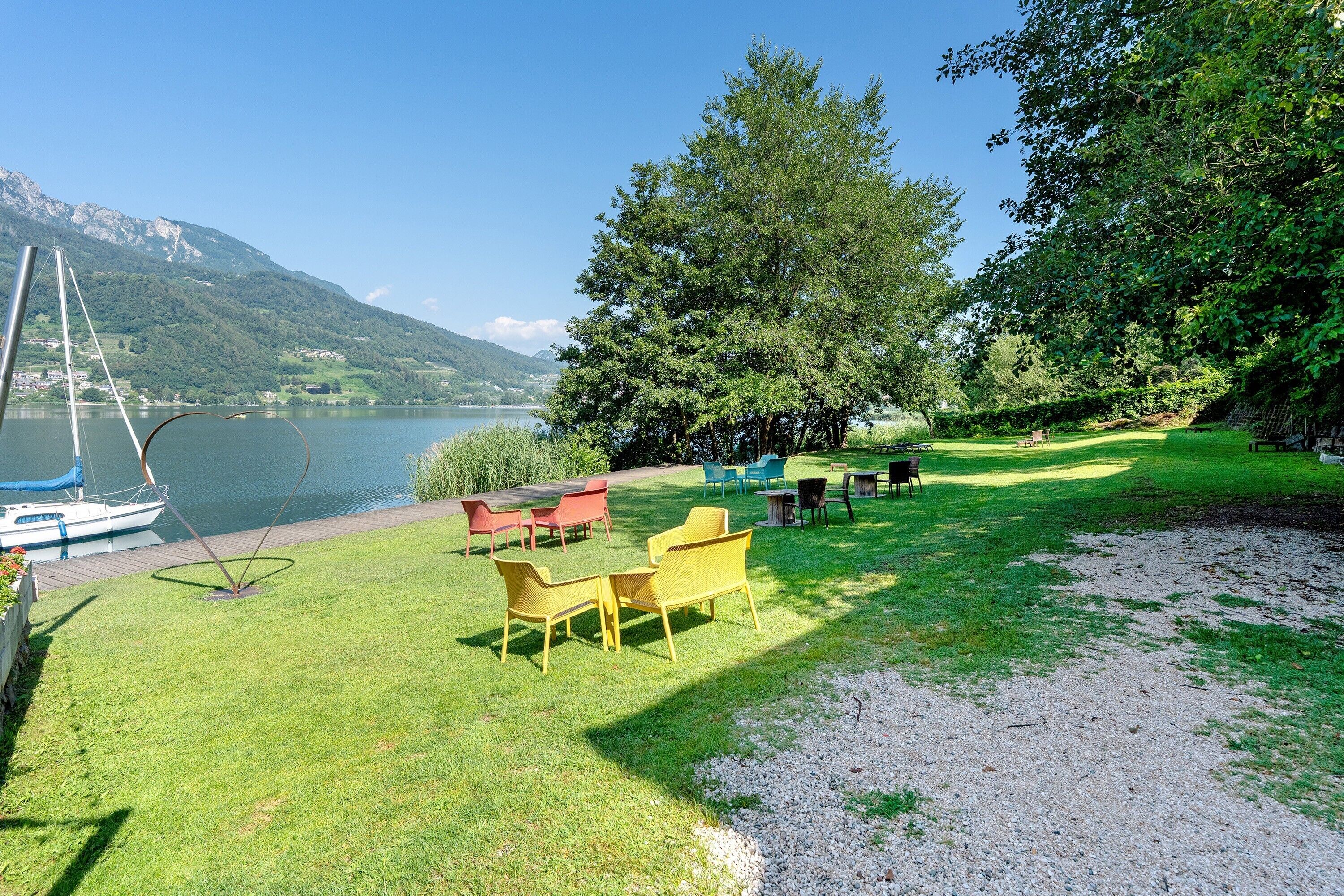 Un espace extérieur aménagé au bord du lac, avec des tables et des chaises, un bateau amarré sur l’eau et une chaîne de montagnes en arrière-plan.