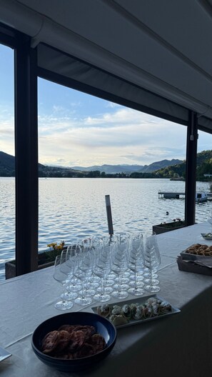 Ein Tisch mit Weingläsern und einer Schüssel Essen, mit Blick auf einen See mit Booten und Hügeln.