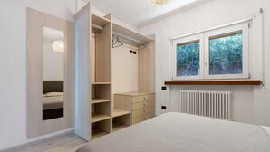 A bedroom with a bed, a wardrobe with open doors, a dresser, and a window with a view of greenery. - Apartment 'Valsugana D’incanto' with Lake View, Private Terrace and Shared Garden (Pergine Valsugana)