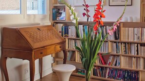 A room with a wooden desk, a white pedestal table, a bookshelf filled with books, a vase with flowers, and a framed picture on the wall. - Spacious, bright apartment near Montmartre! Ideal for families. (Paris)