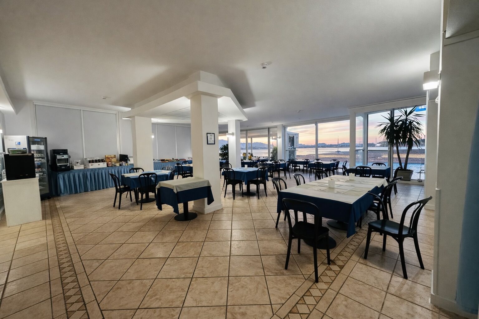 Un vaste espace dédié à la salle à manger, avec des tables et des chaises, de grandes fenêtres offrant une vue dégagée et un sol carrelé.