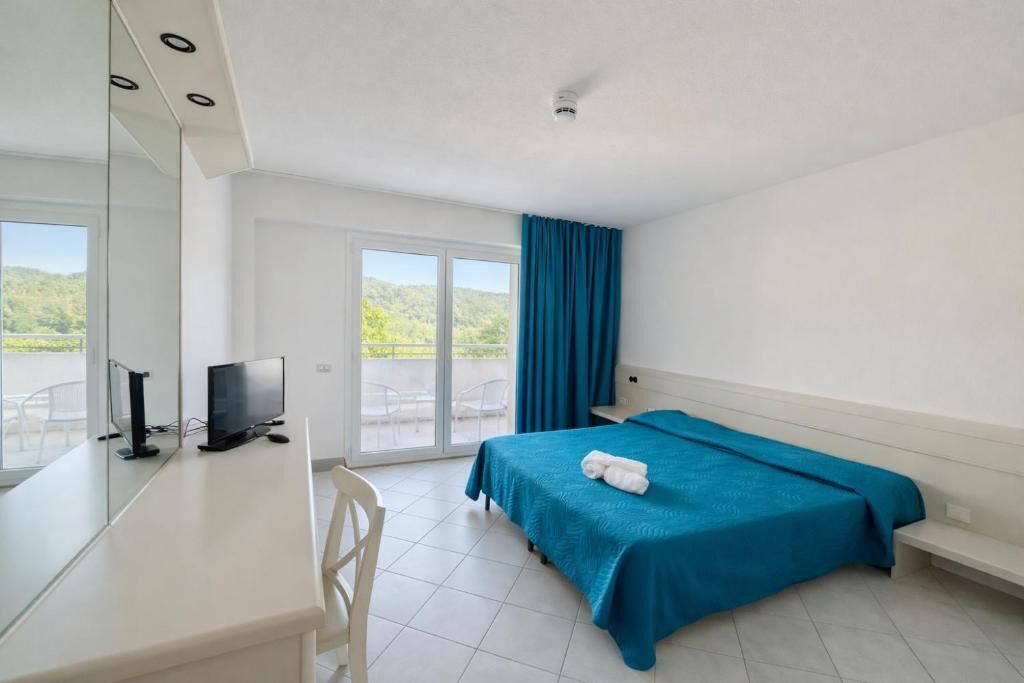A hotel room with a bed, a desk with a TV, a window with curtains, and a view of greenery outside.