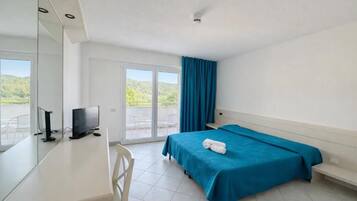 A hotel room with a bed, a desk with a TV, a window with curtains, and a view of greenery outside.