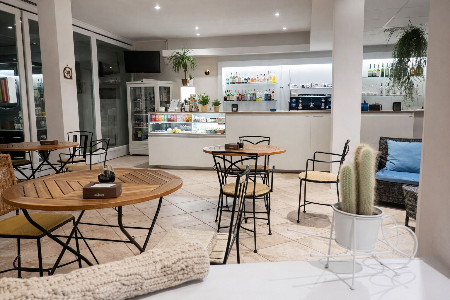 Un café moderno con mesas redondas de madera y sillas de metal negro, una zona de bar con estantes llenos de botellas y una planta de cactus sobre una mesa blanca.