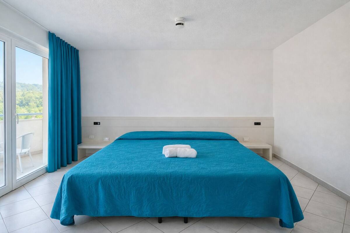 A hotel room with a large bed, a window with curtains, a ceiling light, and a view of greenery outside.
