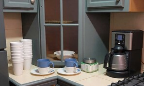 Una cocina con tazas blancas apiladas, tazas azules sobre platillos, una cafetera y una ventana con vistas.