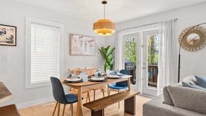 A dining area with a wooden table, chairs, and a bench. There is a large window with blinds, a potted plant, and a painting on the wall.