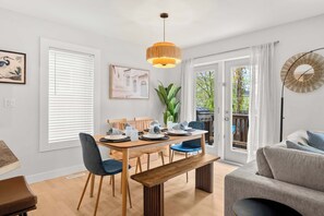 Et spiseområde med et træbord, stole og en bænk. Der er et stort vindue med persienner, en potteplante og et billede på væggen.