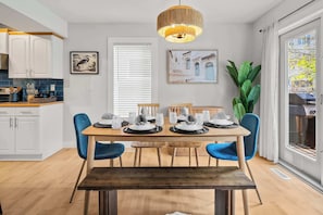 Un coin repas avec une table en bois dressée pour quatre personnes, des chaises bleues, un luminaire suspendu et une grande fenêtre avec des stores.