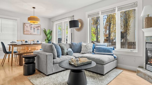 A modern living room with a sectional sofa, a round coffee table, a dining area with a wooden table and chairs, and large windows with a view of trees.