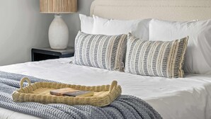 A neatly made bed with white bedding, a woven basket with a book, and a lamp on a bedside table.