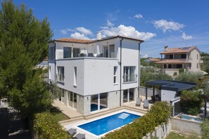 Ein modernes, zweistöckiges Haus mit Swimmingpool, großen Fenstern und einer überdachten Terrasse.
