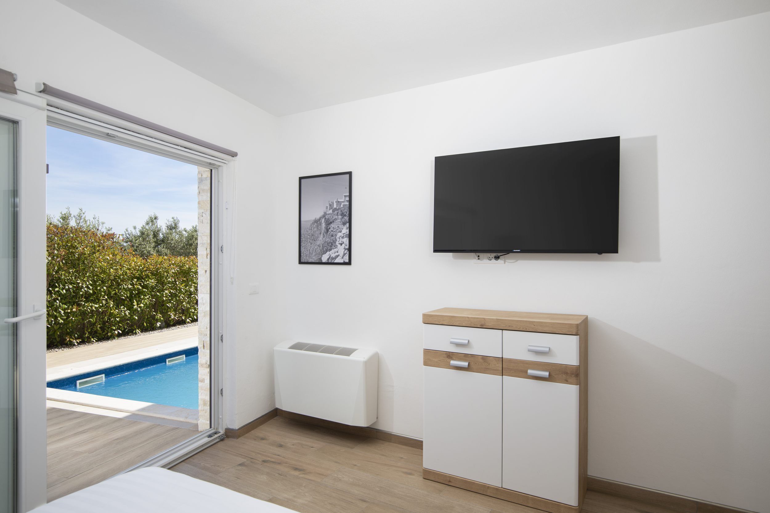Un dormitorio con puerta corrediza al jardín y piscina, un armario blanco, televisor montado y un cuadro en la pared.