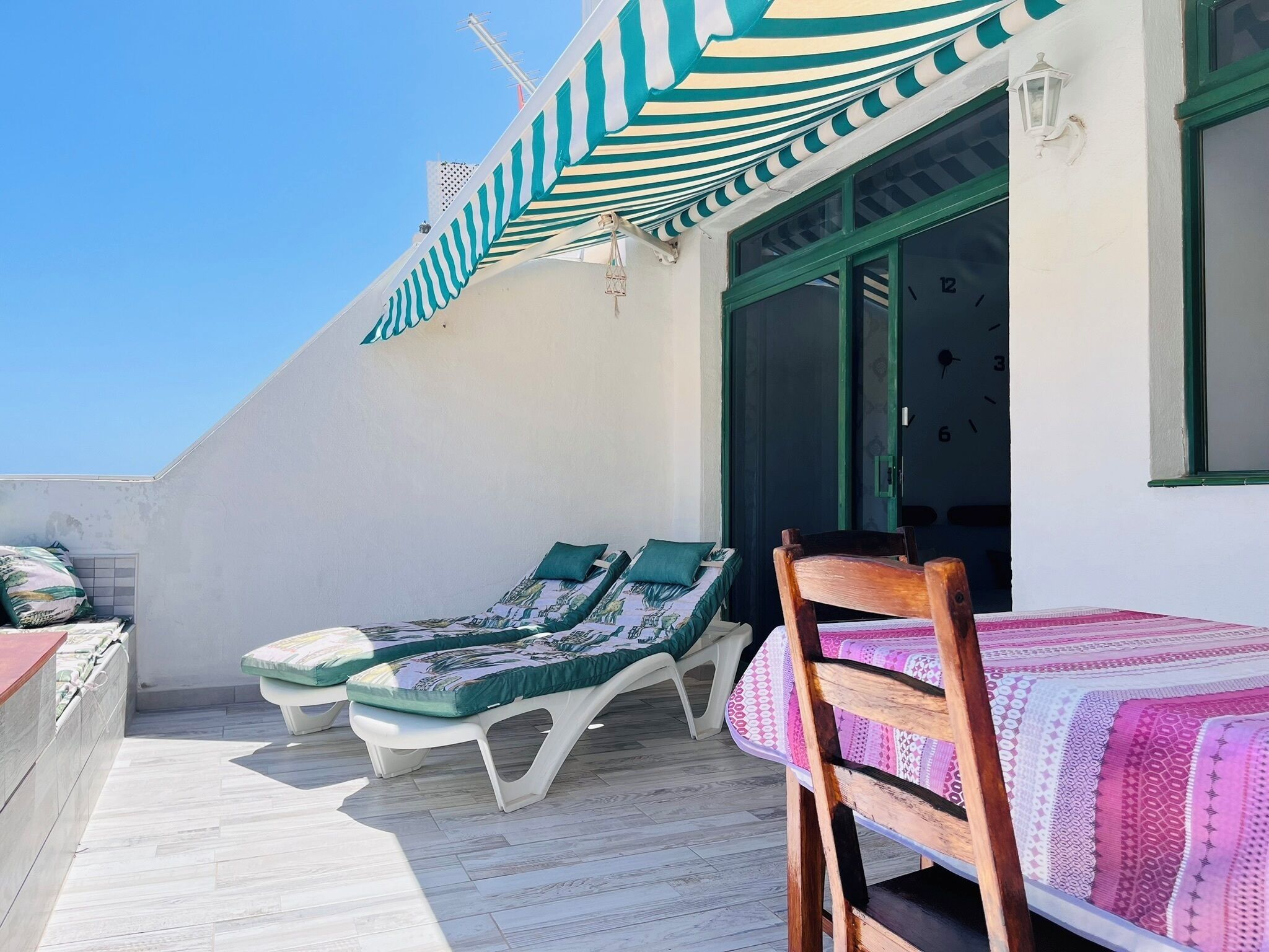 A sunlit outdoor seating area with two lounge chairs and a wooden chair, a striped awning, and a white wall with a window.