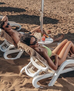 Deux personnes se détendent sur des chaises de plage blanches, une table est placée entre elles, ainsi qu’un parasol de plage et un panier en osier.