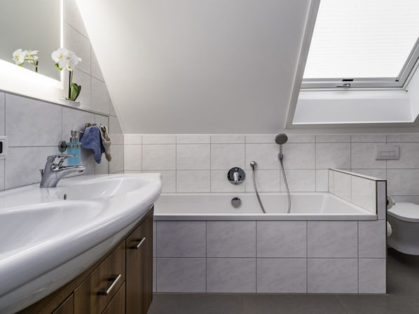 A modern bathroom with a skylight, two sinks, a bathtub, and a toilet. - Seetraum Berger Mainau (Hagnau)