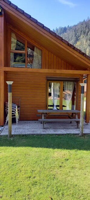 A wooden cabin with a picnic table and chairs, surrounded by a grassy area and a forested mountain backdrop. - Myosotis 2 - Cosy chalet Gérardmer | Swimming pool, terrace, pets welcome (Gérardmer)