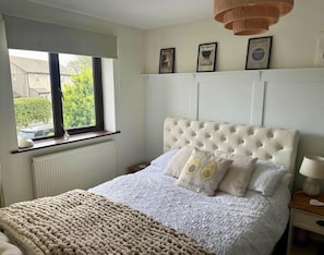 A bedroom with a tufted headboard, a large bed with a white quilt and two decorative pillows, a window with a view of a house and greenery, and a radiator below the window.