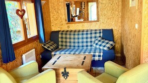 Ein gemütlicher Raum mit Holzbalkendecke, einer Couch mit Karokissen, zwei grünen Sesseln, einem Holztisch und einem Fenster mit Vorhängen. An der Wand hängt ein Spiegel.