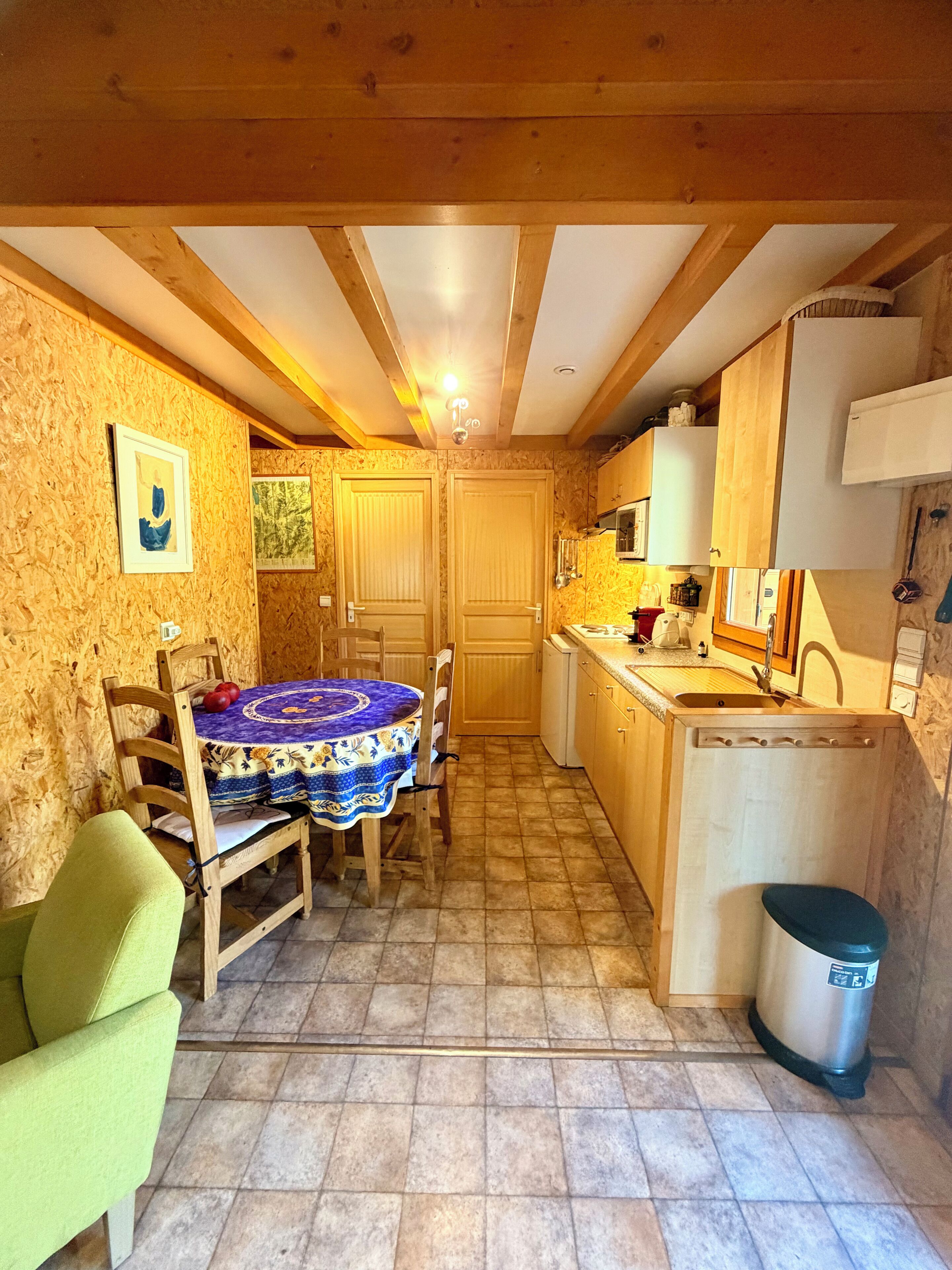 A compact kitchen with wooden cabinets, a dining table with chairs, a trash bin, and a small window.
