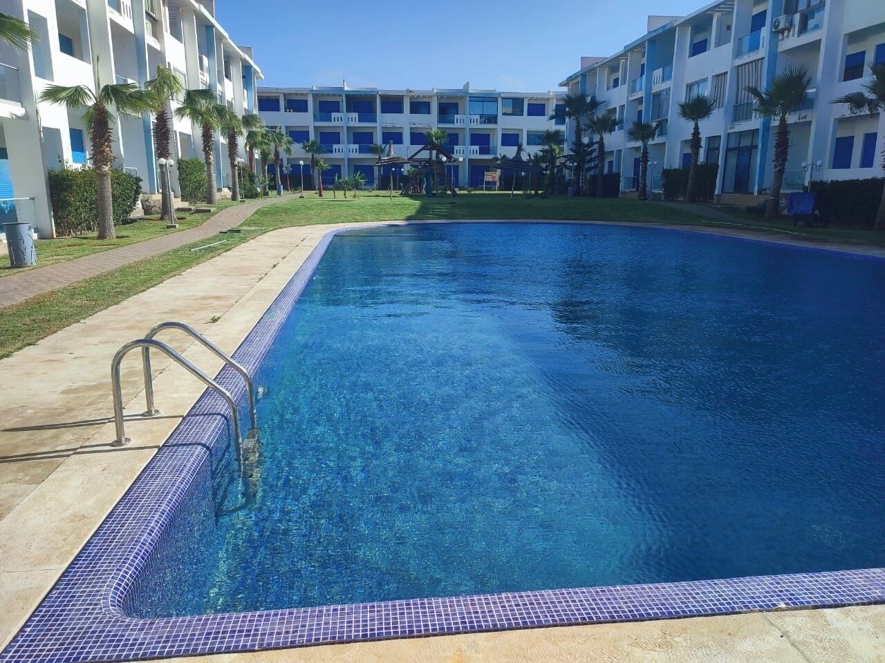 A swimming pool with a tiled edge, surrounded by a building with balconies and palm trees.