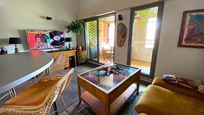 Ein Wohnzimmer mit Glastisch, Korbstühlen, einer Couch, einem Fernseher und einer Pflanze.