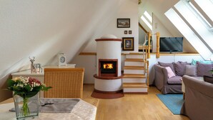 A cozy loft-style room with a fireplace, a sofa, a television, and a wooden staircase. - The vacation apartment in beautiful natural surroundings Relax, unwind and feel good (Hinterzarten)