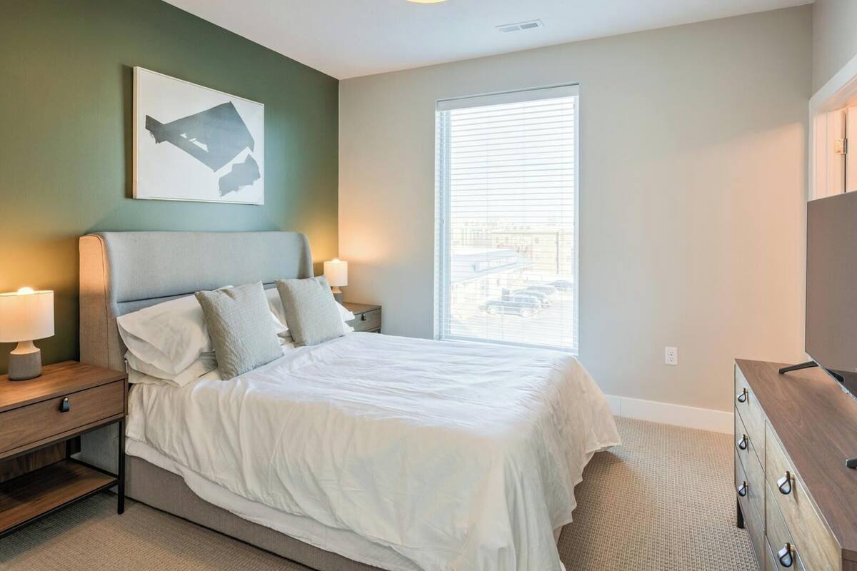 Un dormitorio con cama, lámparas de noche, cómoda, ventana con persianas y un cuadro en la pared.