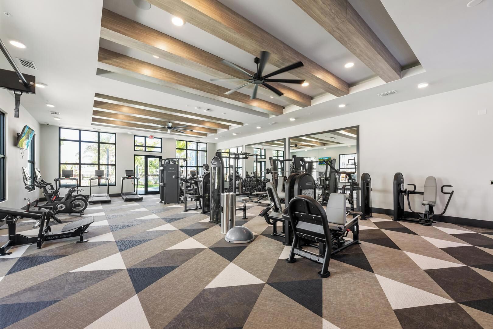 Un gimnasio con diverso equipo de ejercicio, amplias ventanas y piso a cuadros.