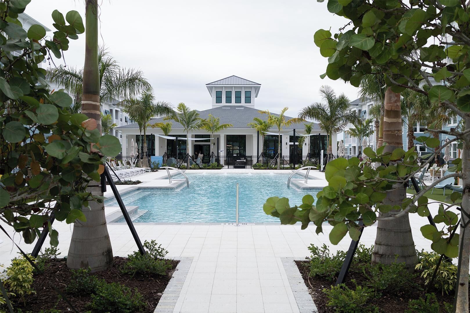 A pool area with a white building in the background, surrounded by palm trees and a tiled walkway.
