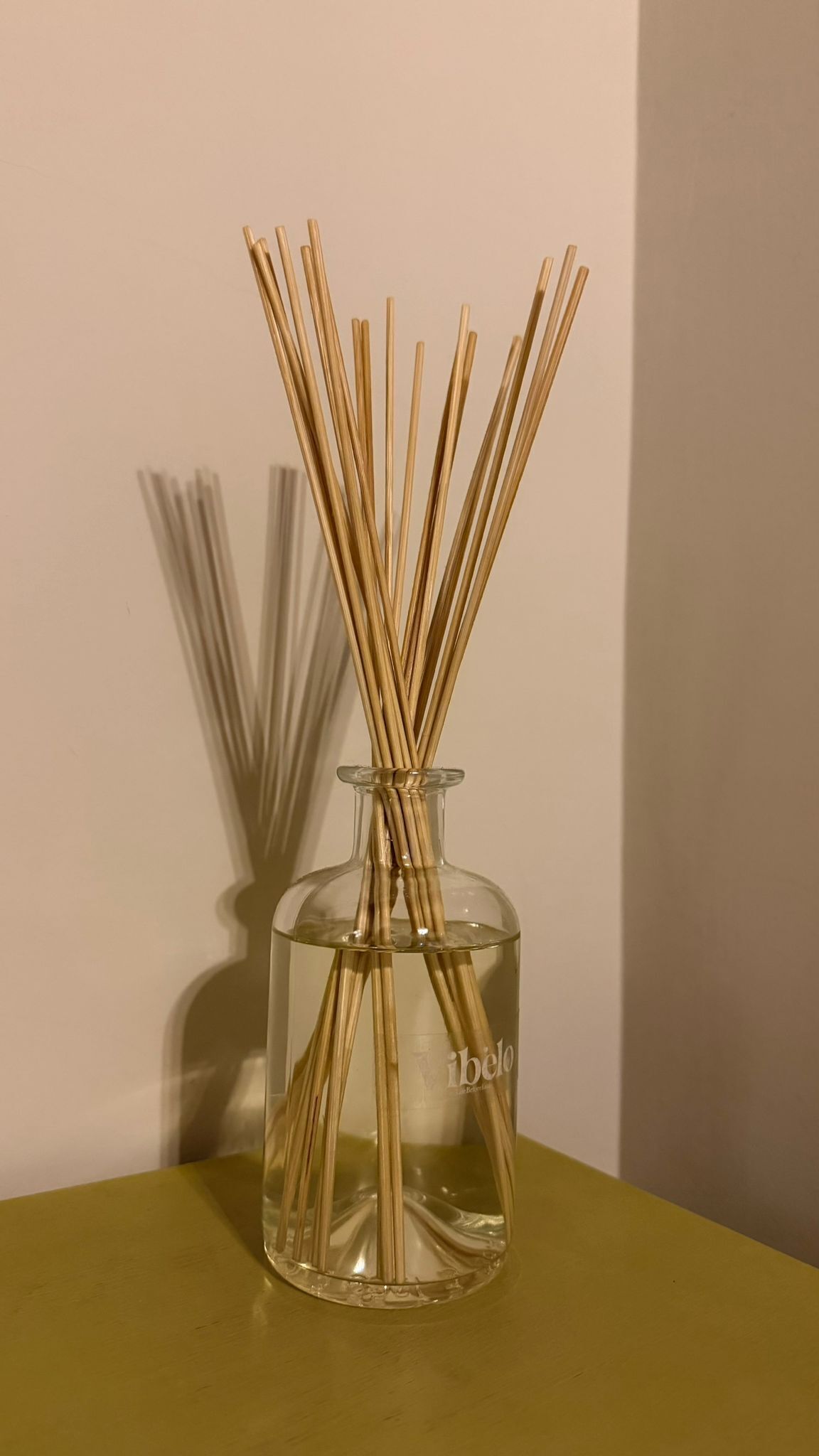 Un vase en verre transparent contenant des bâtonnets en bois, portant l’étiquette « ibélo », est posé sur une surface jaune devant un mur uni.