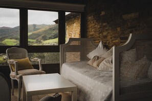 A room with a bed, a chair, and a table, with a view of a landscape through large windows.