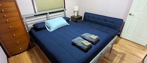 A bedroom with a bed, a wooden dresser, a nightstand, and a window with blinds.