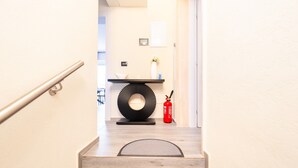 Un escalier mène à une pièce au design moderne, comprenant une décoration murale circulaire noire, un vase blanc avec des fleurs et un extincteur rouge.
