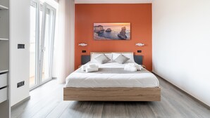Une chambre moderne avec un grand lit, un tableau représentant un coucher de soleil et une vue sur la mer par la fenêtre.