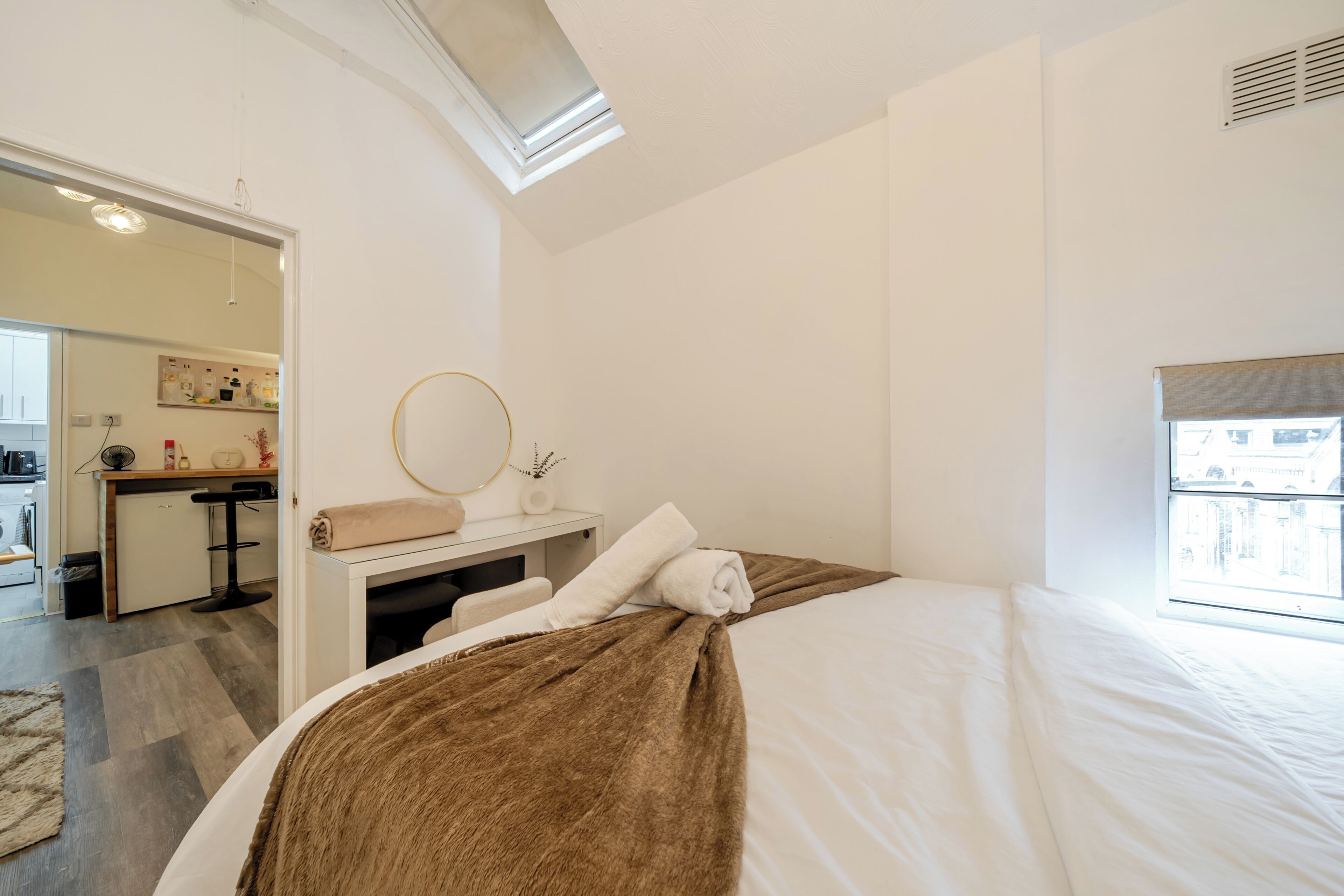 A bedroom with a bed, a bedside table with a mirror, a small desk, and a window with a view.