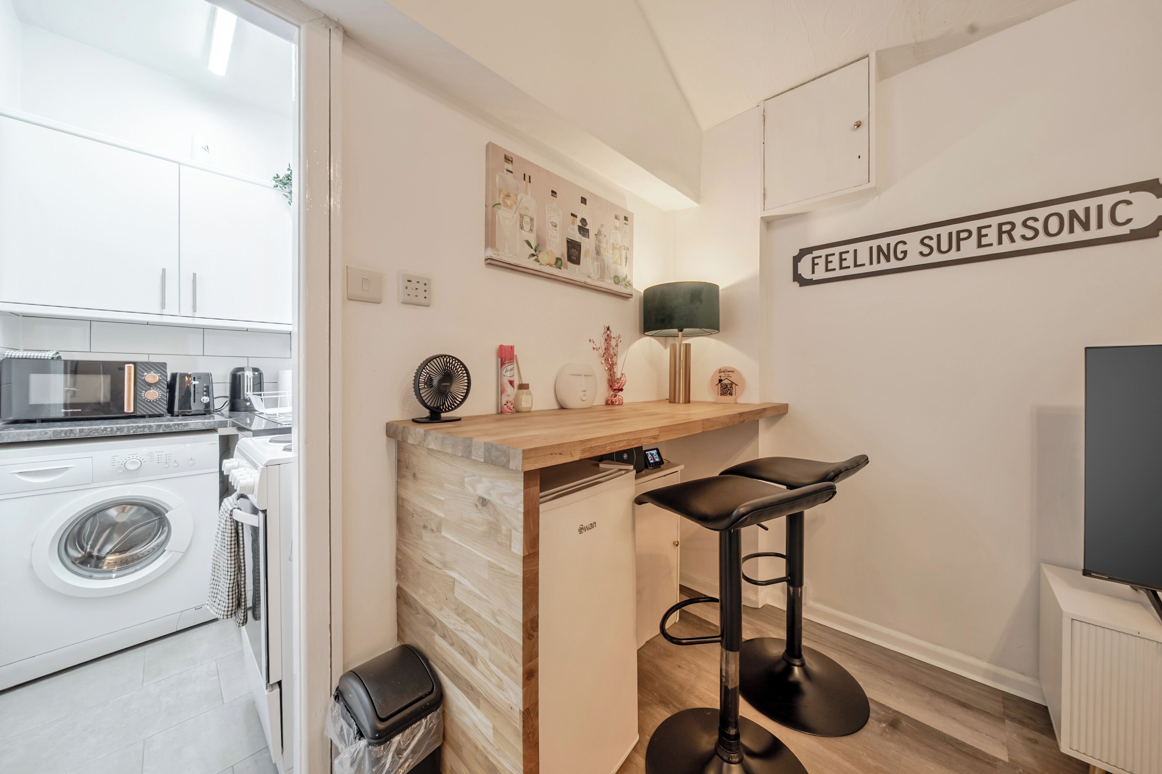 A compact kitchen with a washing machine, a microwave, and a sink. There is a small bar area with stools and a wall-mounted lamp.