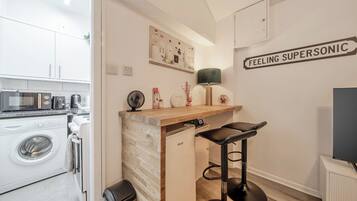 A compact kitchen with a washing machine, a microwave, and a sink. There is a small bar area with stools and a wall-mounted lamp.