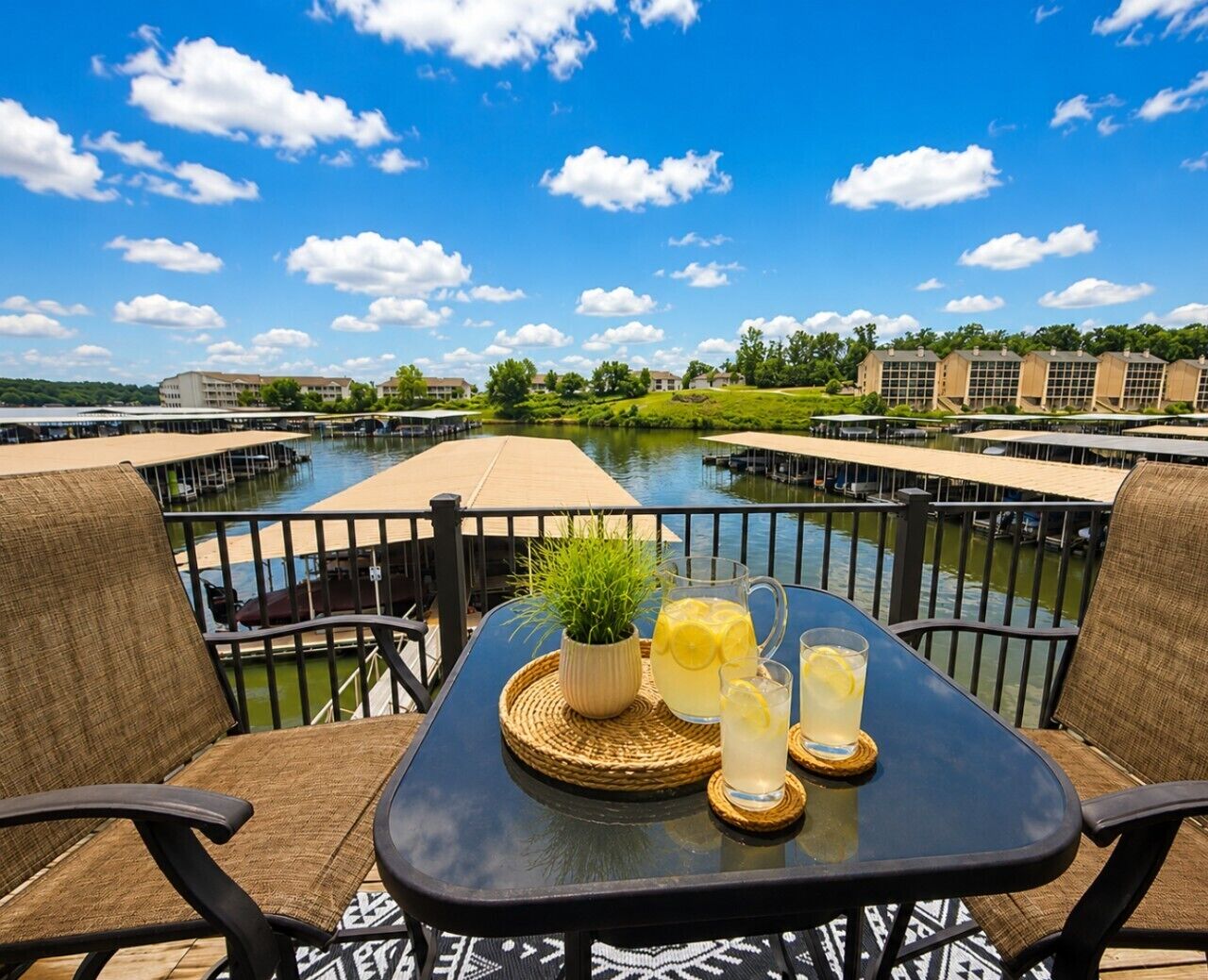 A table with a pitcher, glasses, and a plant on a balcony overlooking a water body with buildings in the background.