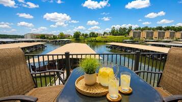 A table with a pitcher, glasses, and a plant on a balcony overlooking a water body with buildings in the background.
