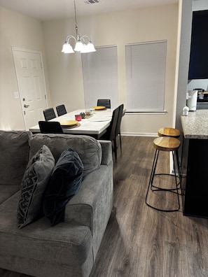 Ein modernes Wohnzimmer mit grauem Sofa, einem Esstisch mit Stühlen, einer Küchenzeile mit Arbeitsplatte und einem hölzernen Hocker.