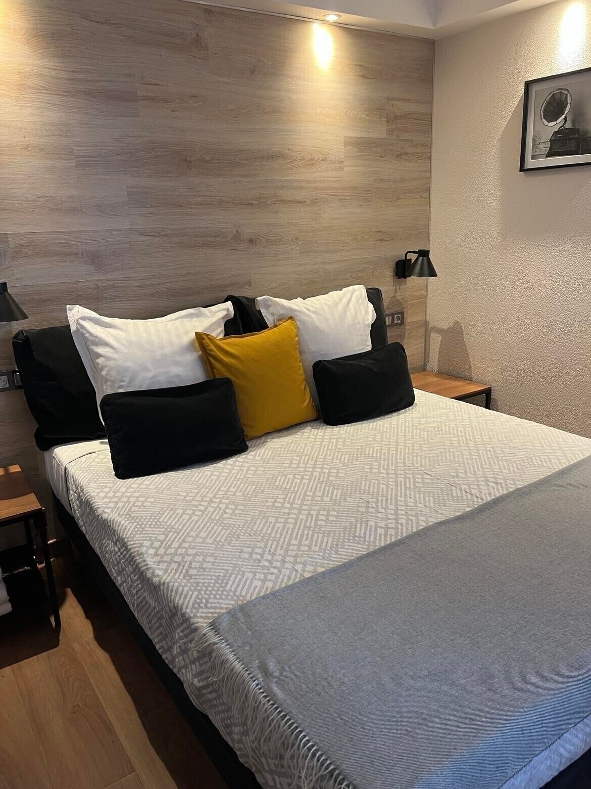 Een netjes opgemaakt bed met een gebedrukte spreideken, diverse kussens en een aan de muur bevestigd fotolijstje.