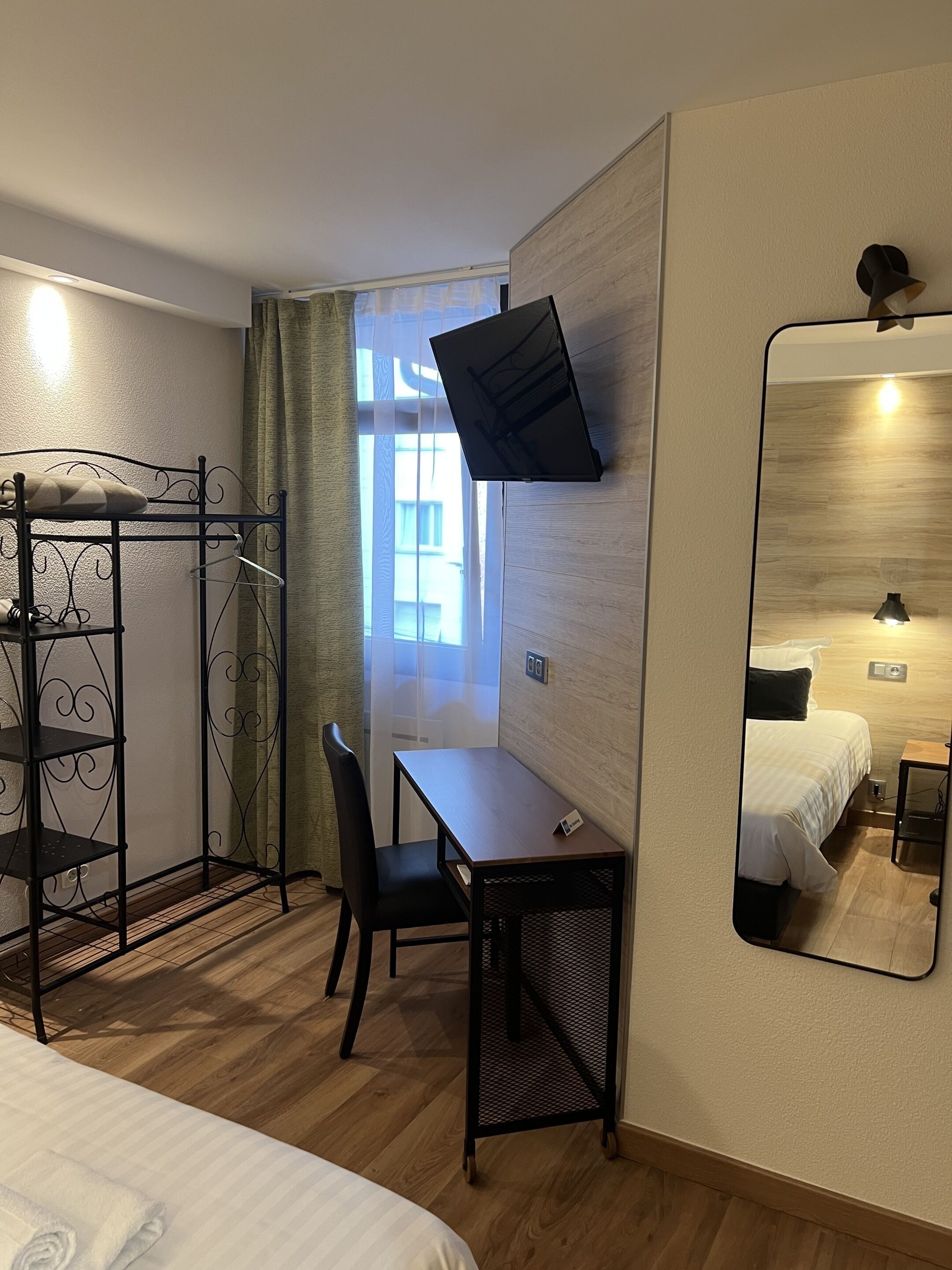 A compact hotel room with a bed, a desk, a chair, a TV mounted on the wall, and a mirror.